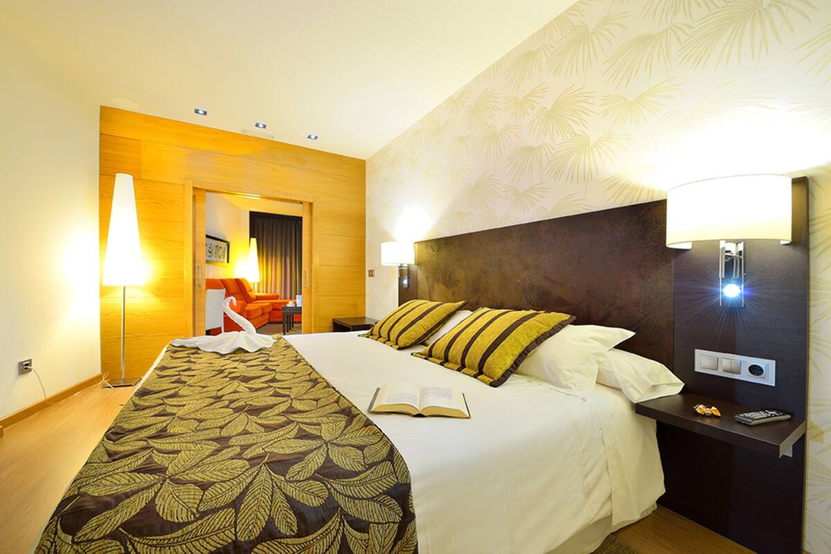 A hotel room with a large bed, a bedside table with a lamp, and a view of the hallway.