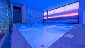 Una piscina interior moderna con vistas al atardecer, paredes con azulejos y zona de baño transparente.
