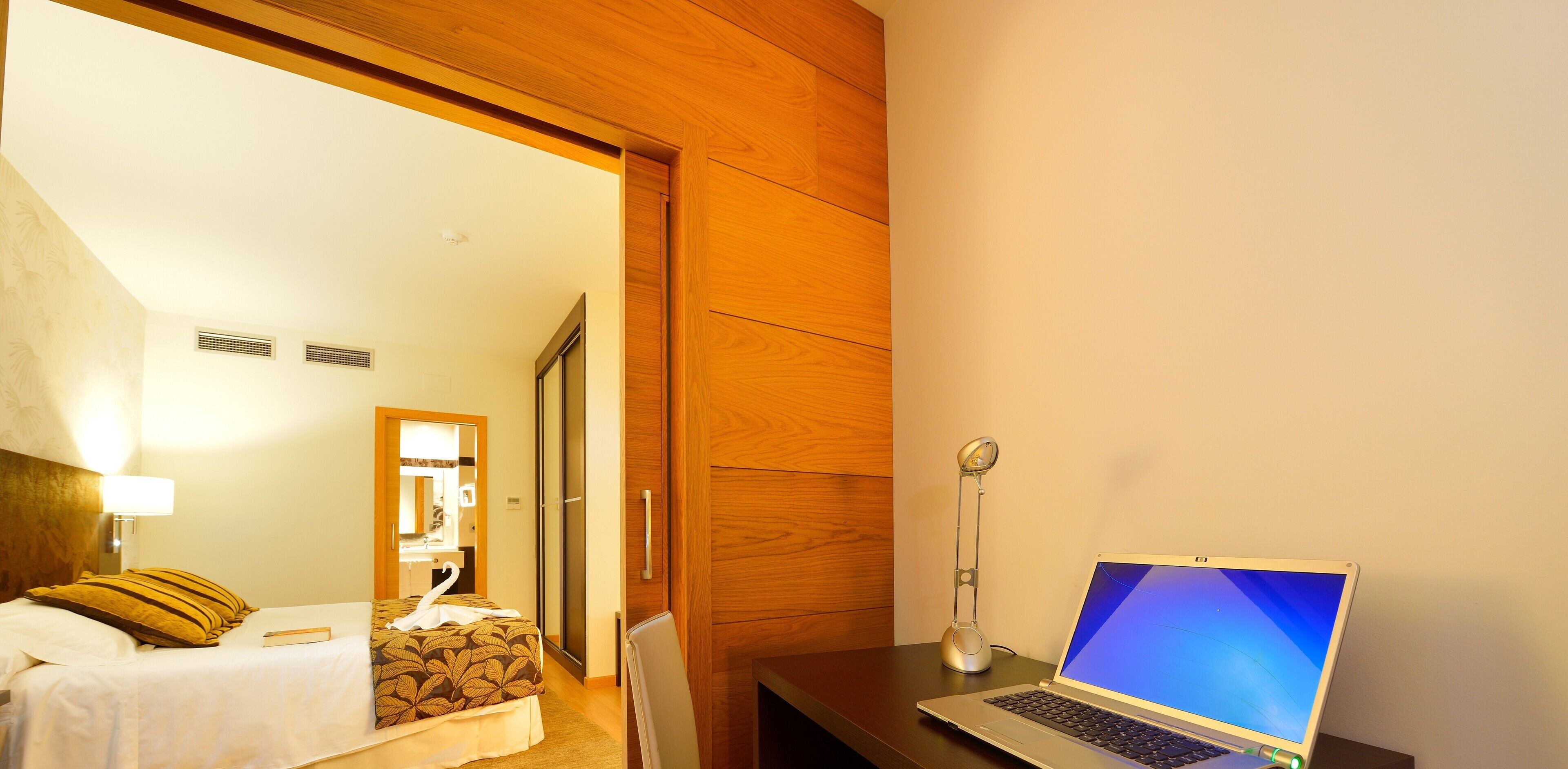 Una habitación de hotel con una cama, un escritorio con una computadora portátil, un espejo y un armario de madera.
