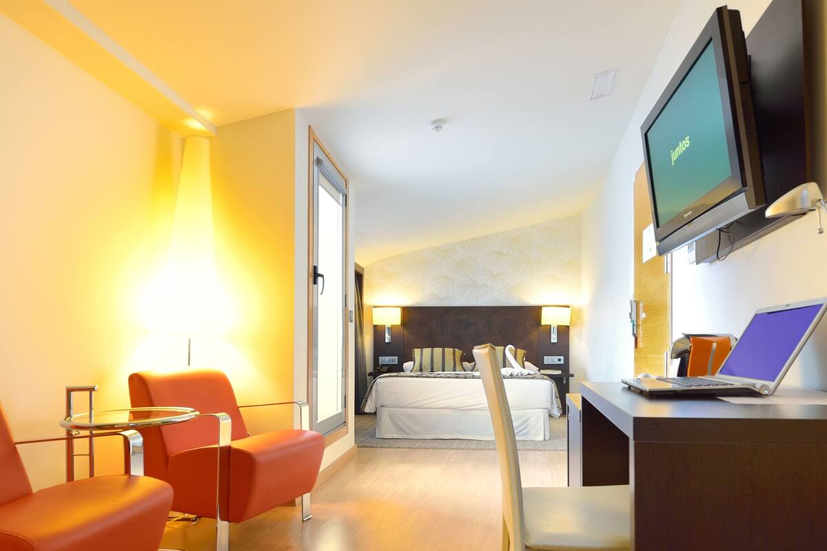 A hotel room with a bed, a desk with a laptop, a TV, and two chairs.