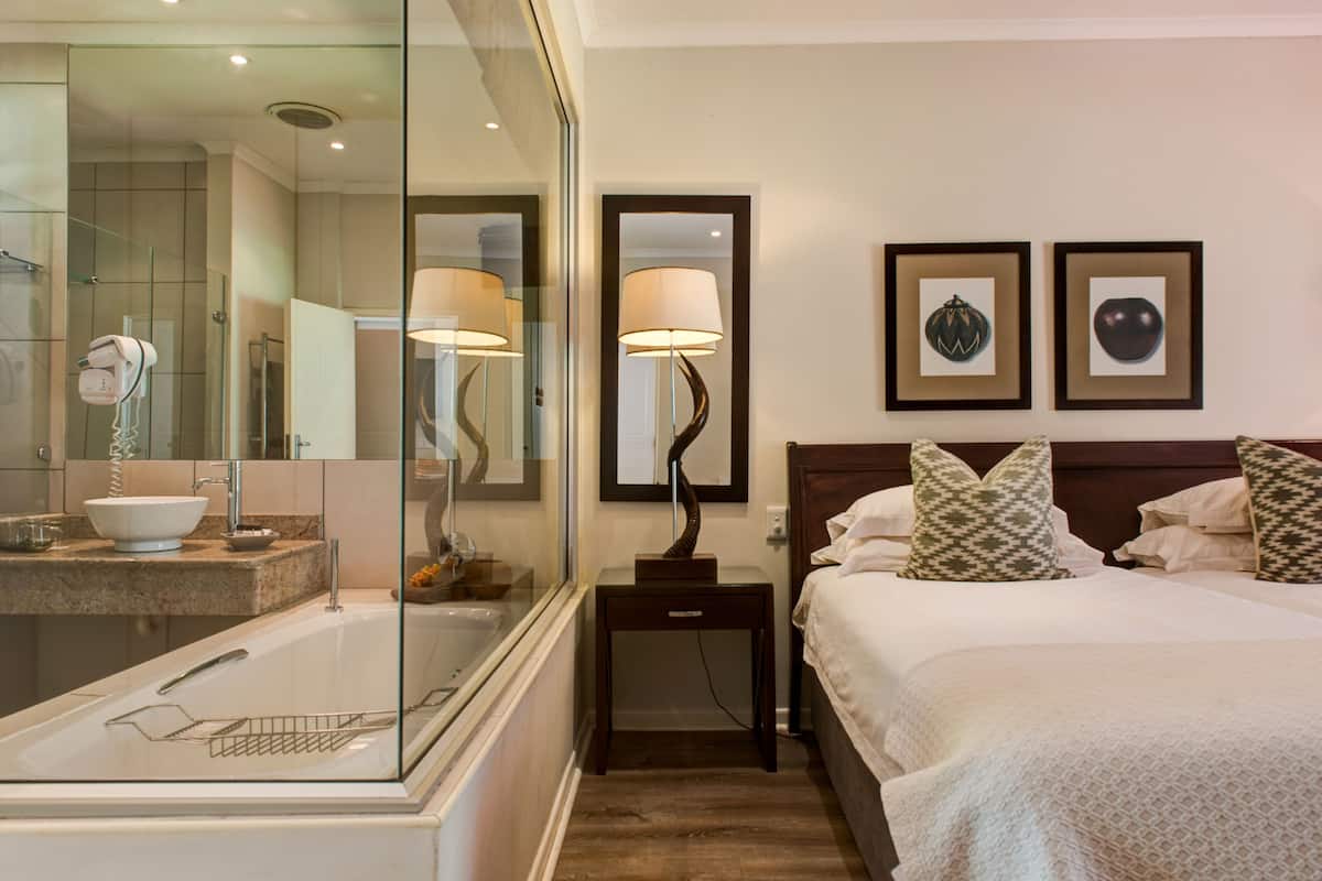 A hotel room with a large mirror, a bathtub, a bedside table with a lamp, a bed with patterned pillows, and two framed pictures on the wall.