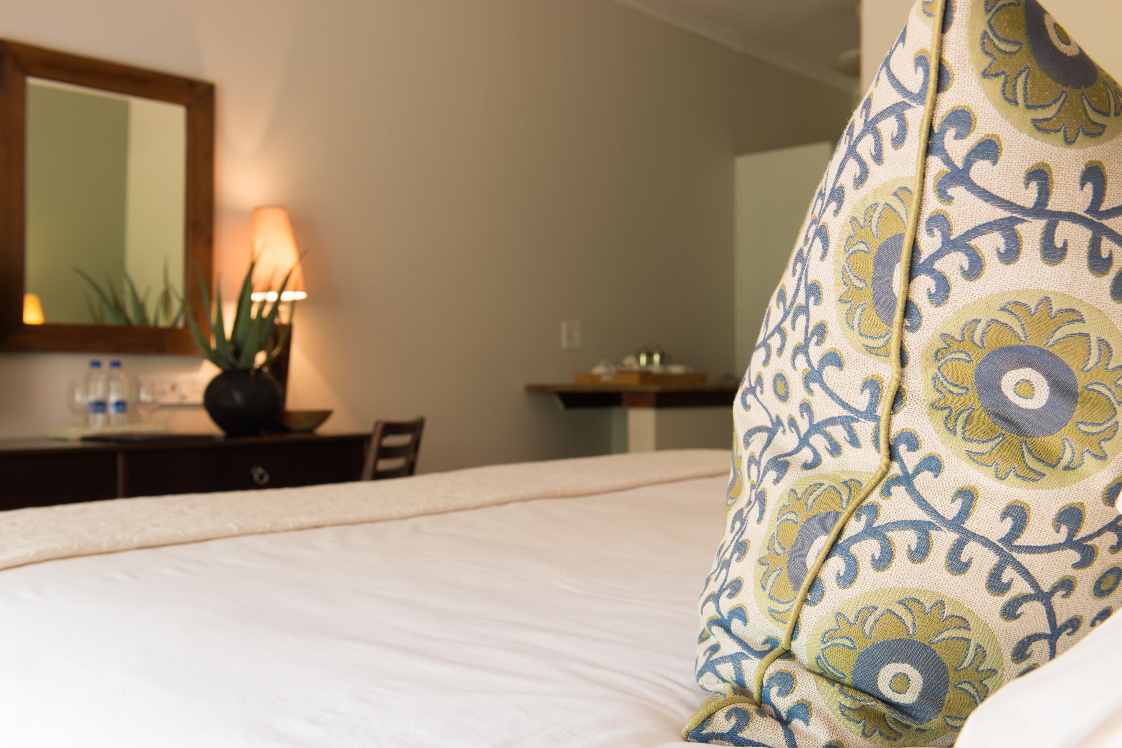 A hotel room with a bed, a desk with a lamp, a mirror, and a plant.