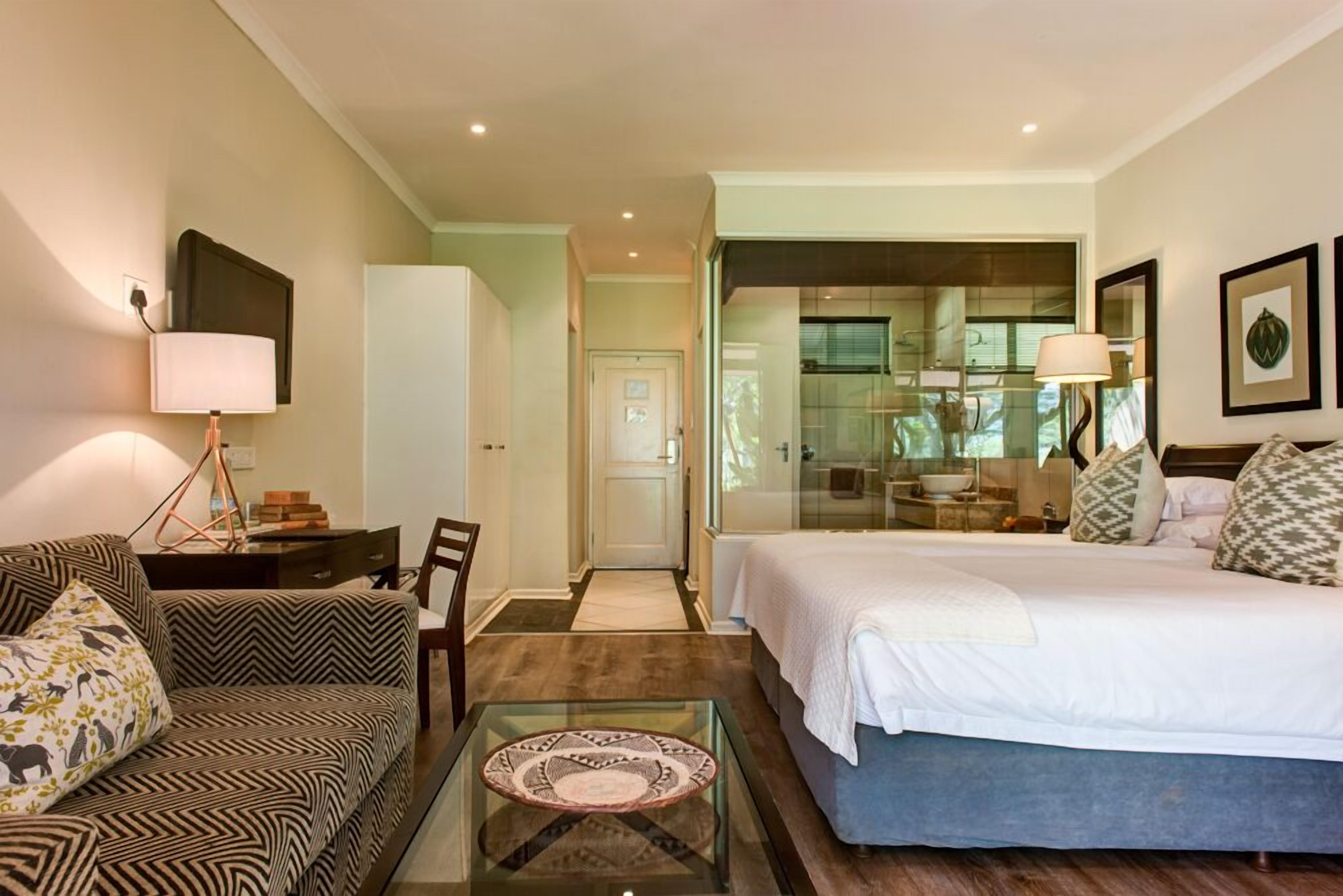 A hotel room with a bed, a sofa, a glass coffee table, a desk, and a chair. There is a kitchen area visible through a glass door.