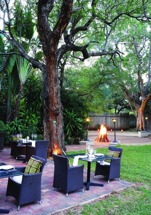 An outdoor dining area with wicker furniture, a fire pit, and lush greenery.
