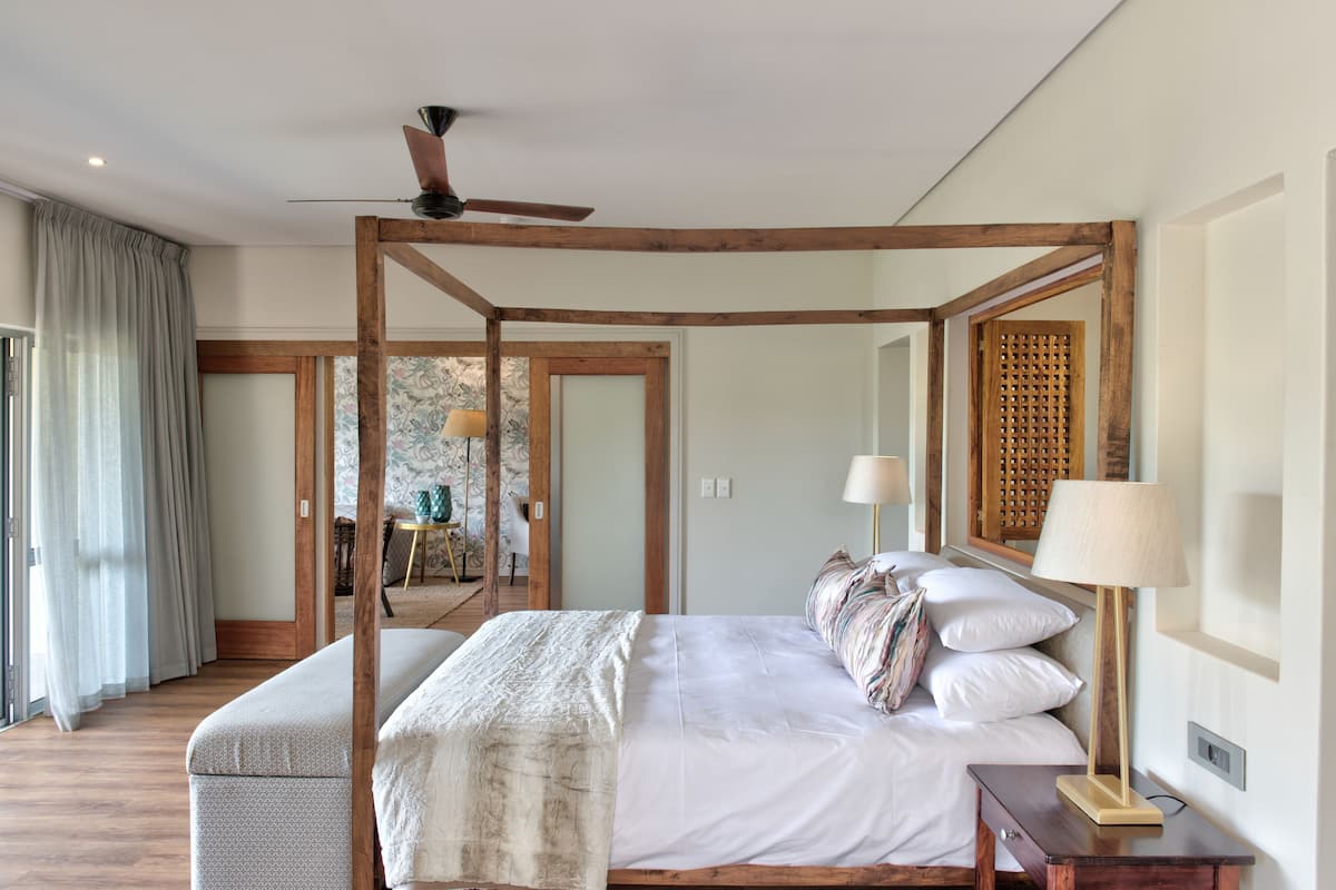 A four-poster bed with a wooden frame, a bedside table with a lamp, and a view of a dining area through a glass door.