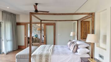 A four-poster bed with a wooden frame, a bedside table with a lamp, and a view of a dining area through a glass door.