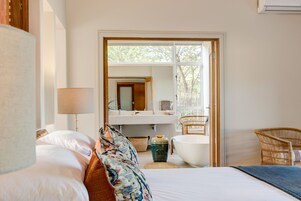 Quarto de hotel com cama, uma janela grande, banheiro com banheira e uma cadeira de vime.