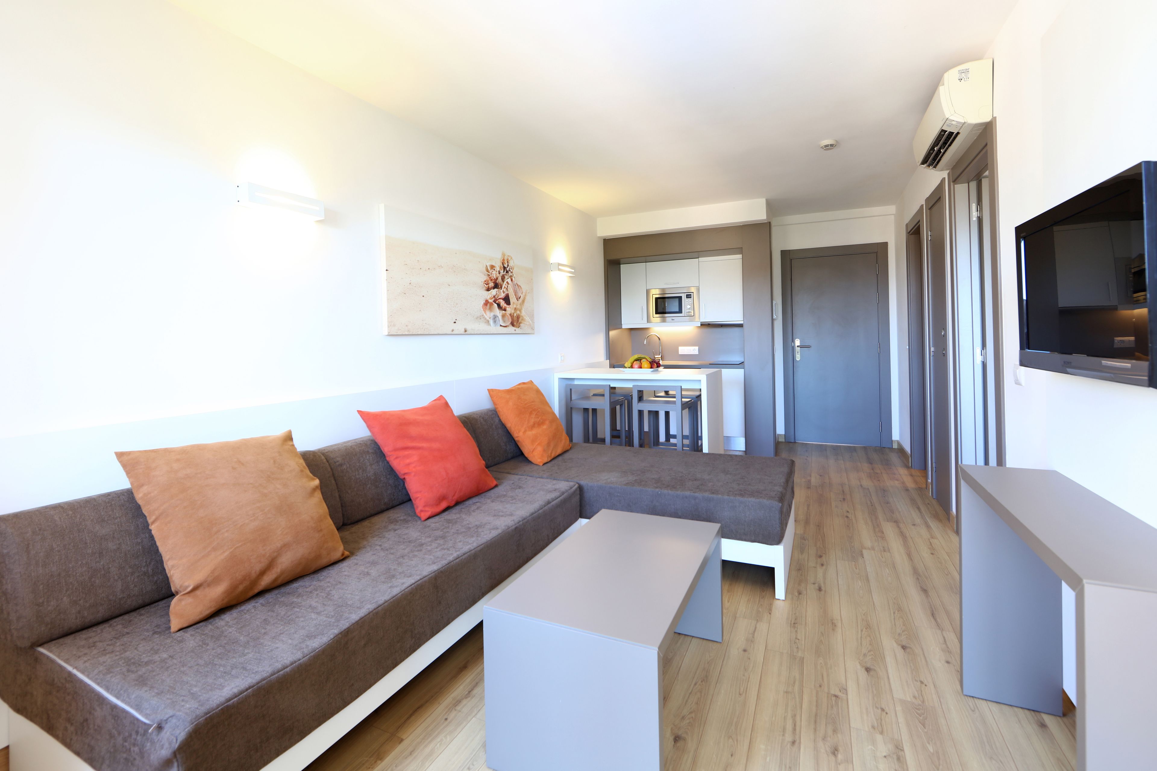 A modern living room with a grey sectional sofa, a white coffee table, and a kitchen area with a microwave and sink.