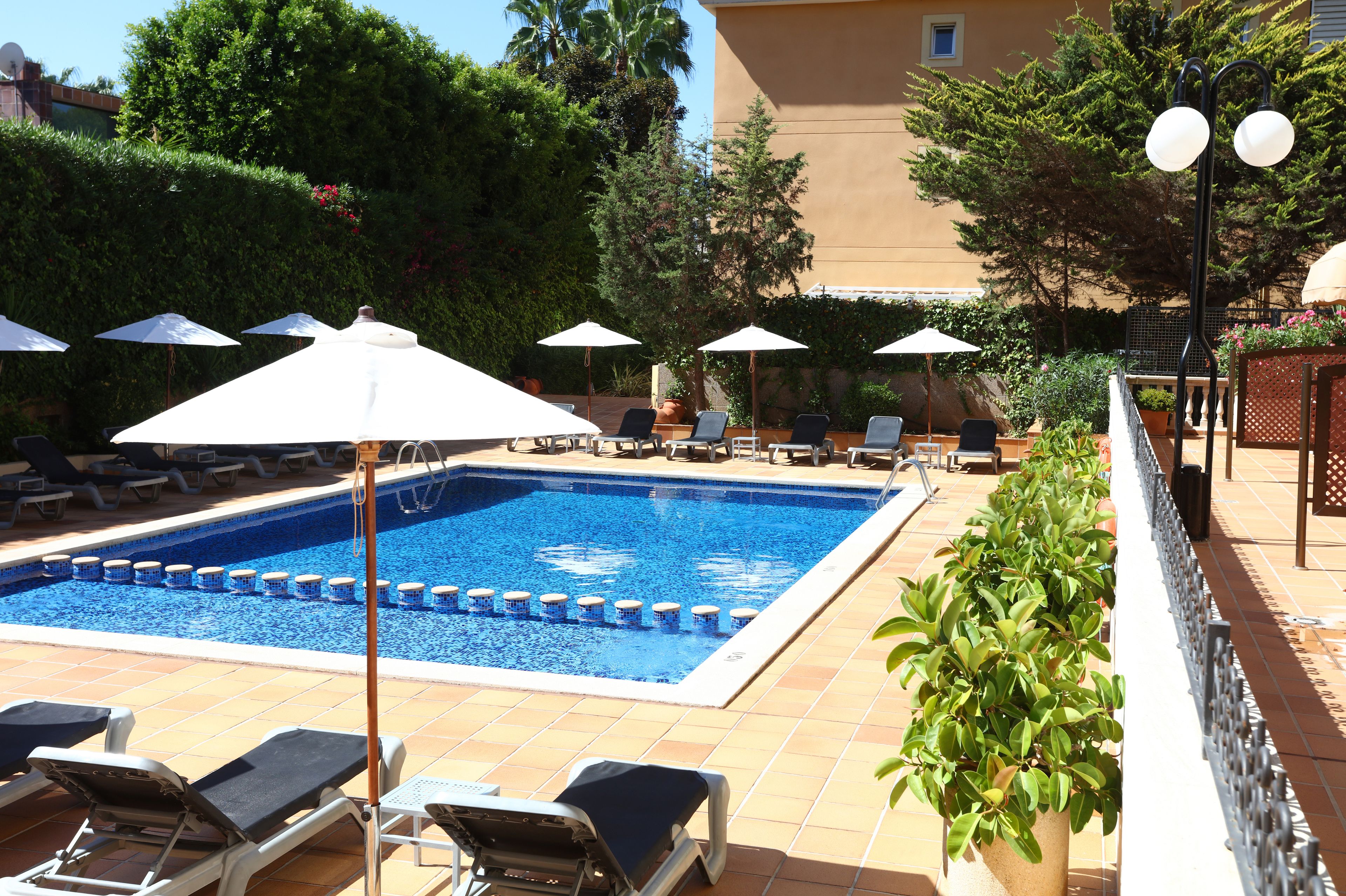 Piscina con sillones de descanso y sombrillas, rodeada de vegetación y un edificio.