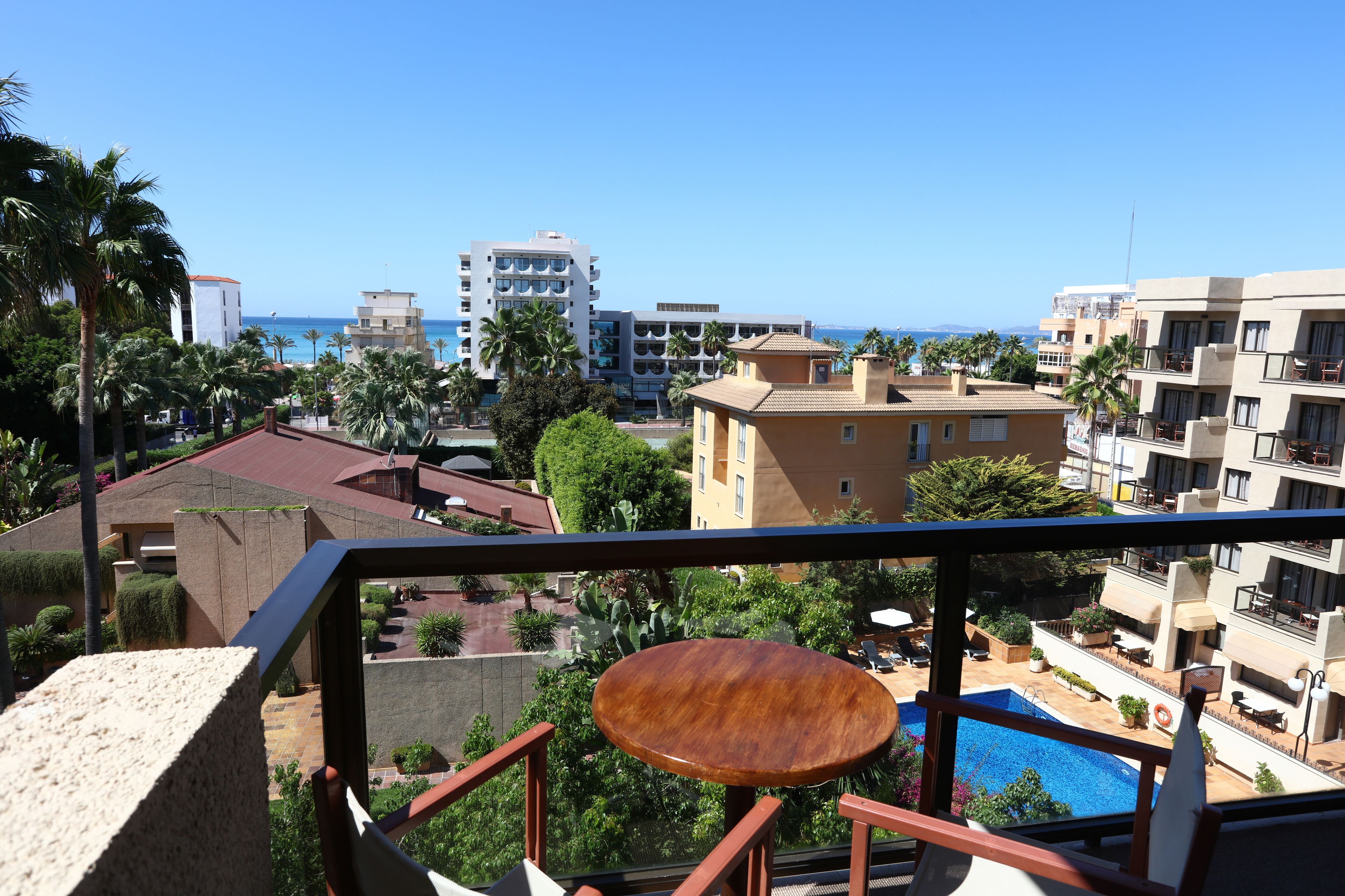 Varanda com mesa de madeira, vista para piscina e para o mar e edifícios ao redor.