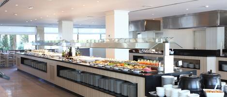 Buffet montado com diversos alimentos, um balcão com pratos e uma área de cozinha moderna ao fundo.