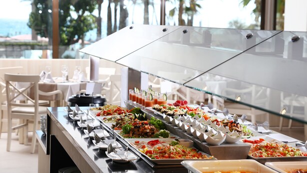 A buffet with various food options, including salads, meats, and desserts, in a bright room with large windows and a view of palm trees. - Apartamentos Jade - Adults Only +16 (Palma de Mallorca)
