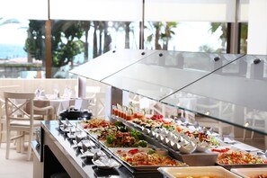 Buffet com diversas opções de comida, incluindo saladas, carnes e sobremesas, em um espaço iluminado com janelas amplas e vista para palmeiras.
