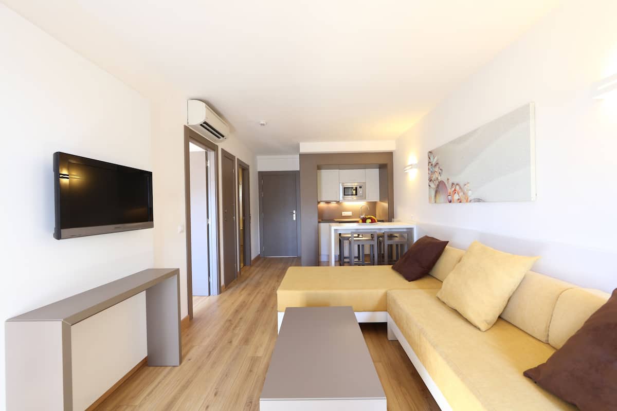 A modern living room with a sofa, a flat-screen TV, and a kitchen area in the background.