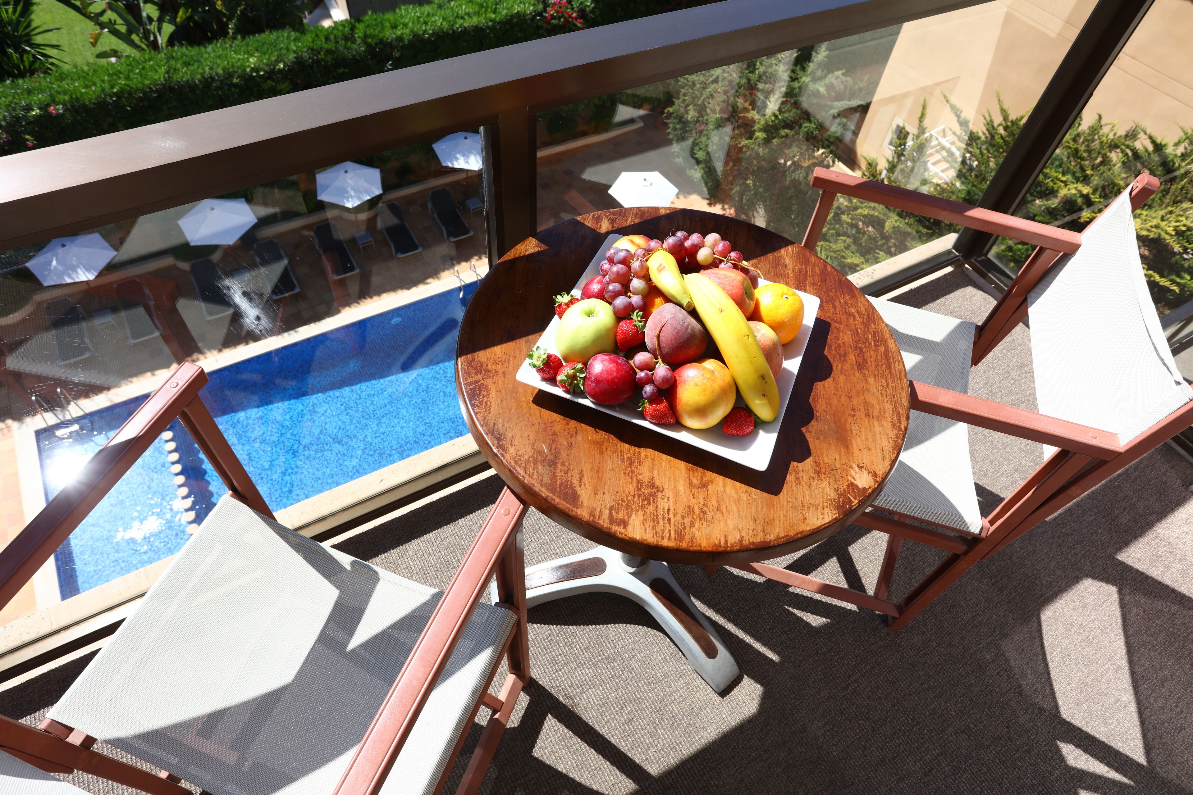 Una mesa redonda de madera con una bandeja de frutas, dos sillas blancas y una barandilla de vidrio con vista a una piscina.
