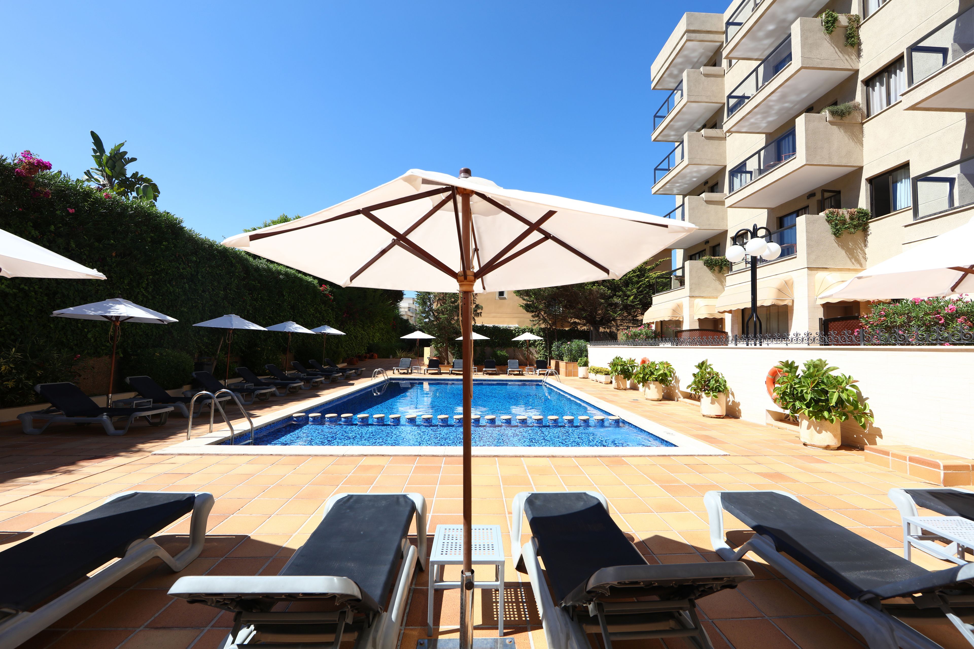 En swimmingpool med liggestole og parasoller, omgivet af en bygning med balkoner og plantekasser.