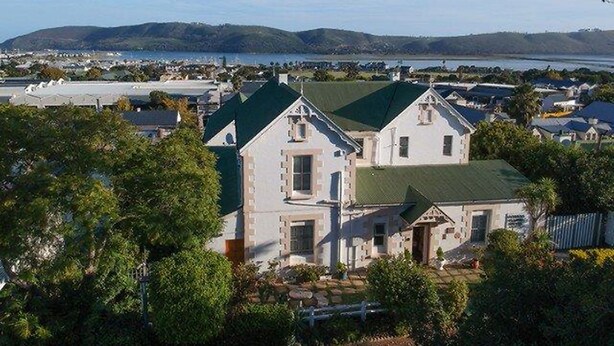 A large, historic house with a green roof, surrounded by lush greenery and a scenic view of a lake and hills. - Knysna Manor House (Knysna)