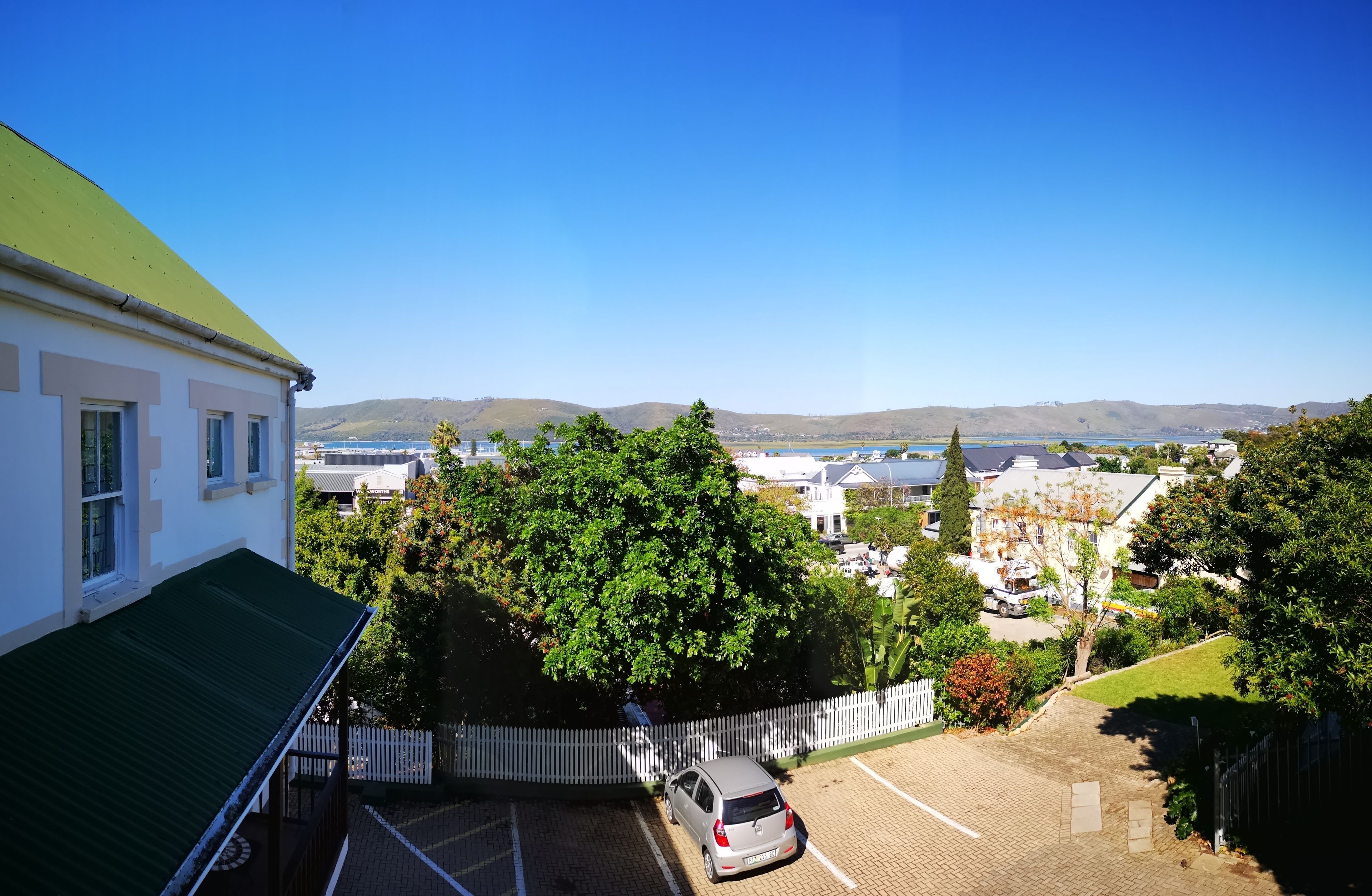 A residential area with a parking lot, a white house with a green roof, and a view of hills and water in the distance.