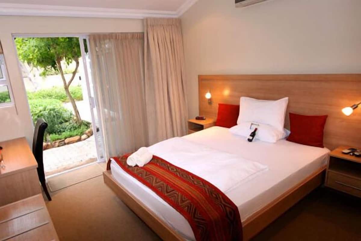 A hotel room with a large bed, a wooden headboard, two bedside tables with lamps, and a view of greenery through the door.