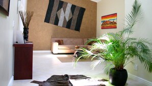 A living room with a sofa, a coffee table, a large potted plant, a rug, and a wall-mounted painting. - Hajo's Lodge & Spa (Cape Town)