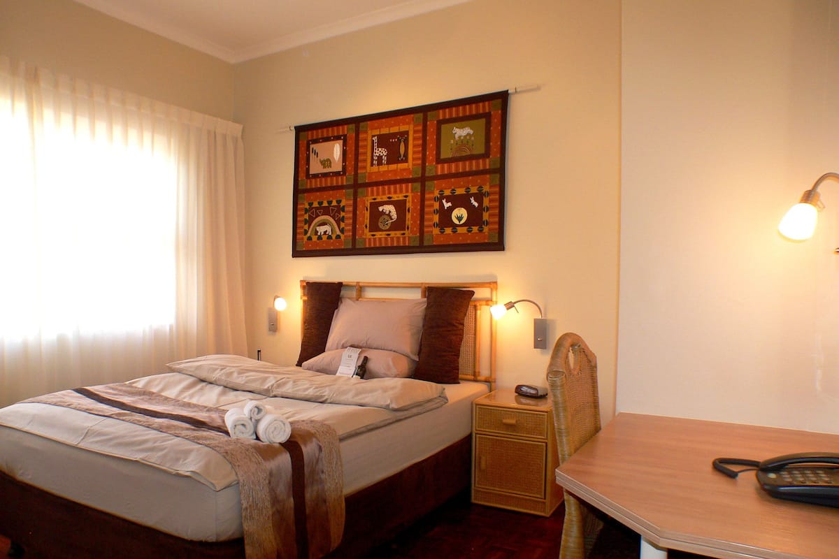 A neatly made bed with a wooden headboard, a bedside table with a lamp, and a wall-mounted quilt with animal prints.