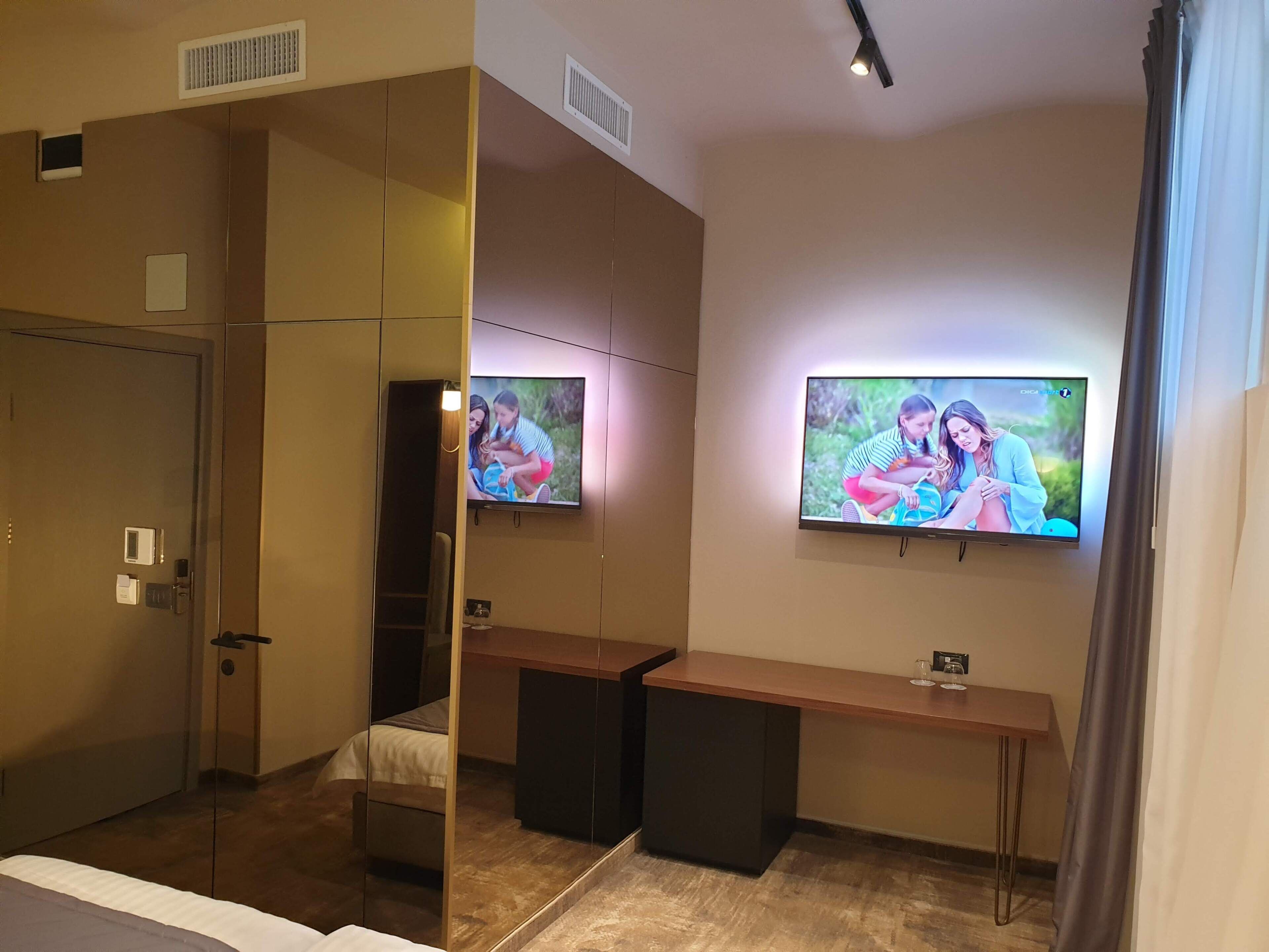 Habitación de hotel con cama, escritorio, dos televisores montados en la pared, un espejo y una puerta.