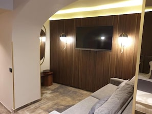 A modern living room with a flat-screen TV mounted on a wooden wall, two wall-mounted lights, a grey sofa, and a small wooden side table.