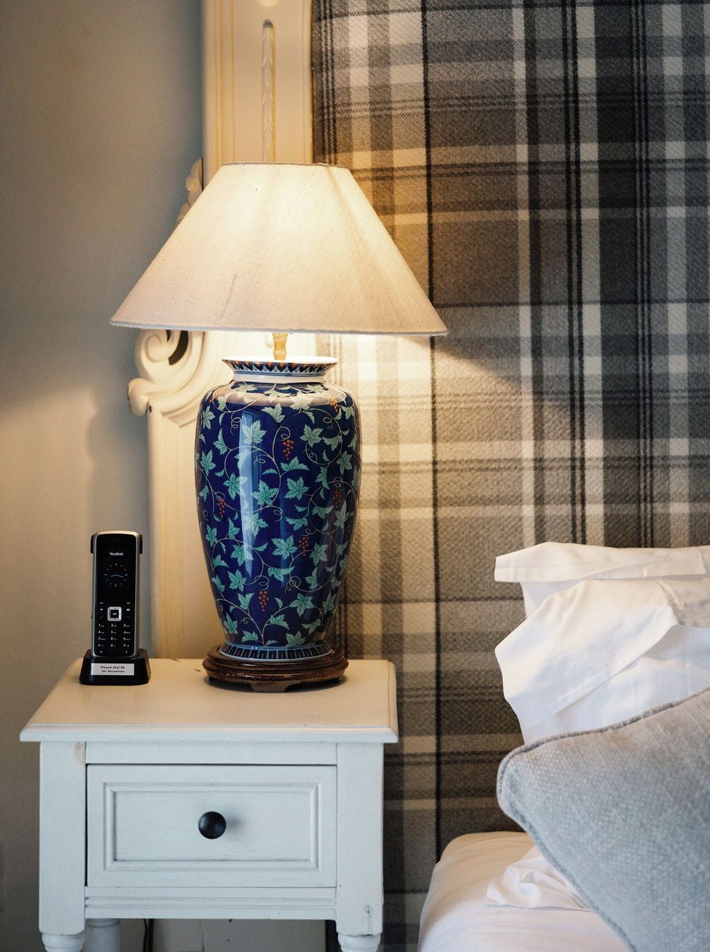 Une table de chevet avec une lampe et une télécommande, une table de nuit blanche et un rideau à carreaux.