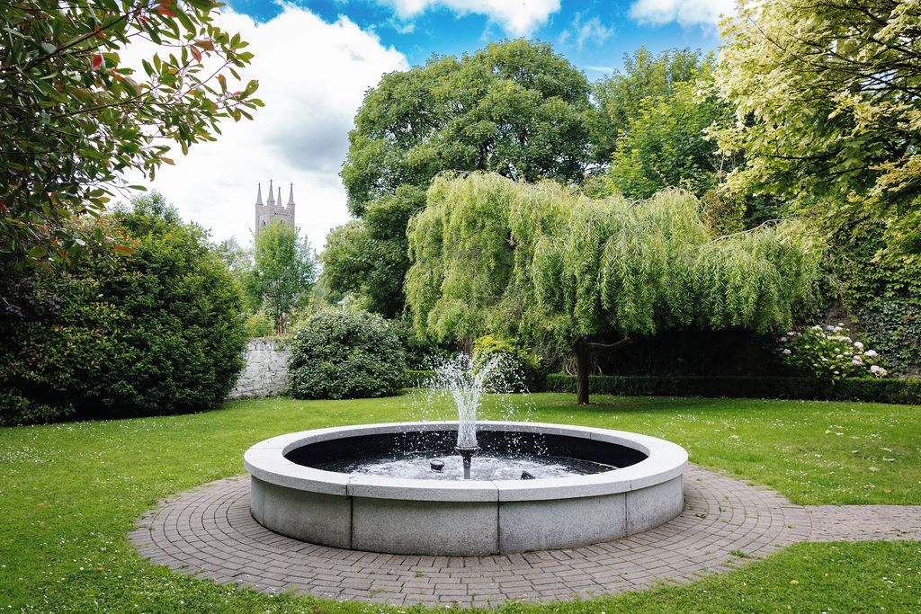 Ein runder Steinbrunnen mit Wasserstrahlen in einem angelegten Garten mit Bäumen und einem Gebäude in der Ferne.