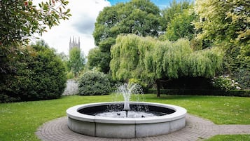 Ein runder Steinbrunnen mit Wasserstrahlen in einem angelegten Garten mit Bäumen und einem Gebäude in der Ferne.