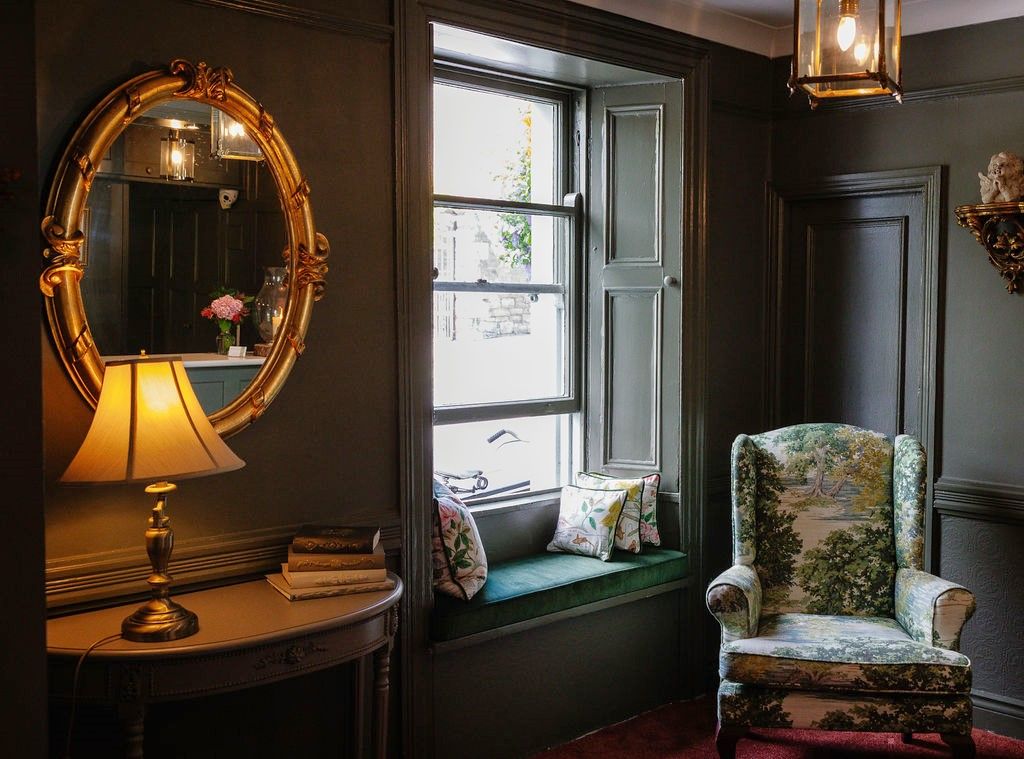 A room with a large mirror, a window seat with cushions, a chair with a floral pattern, and a table with a lamp.