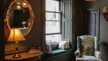 A room with a large mirror, a window seat with cushions, a chair with a floral pattern, and a table with a lamp.