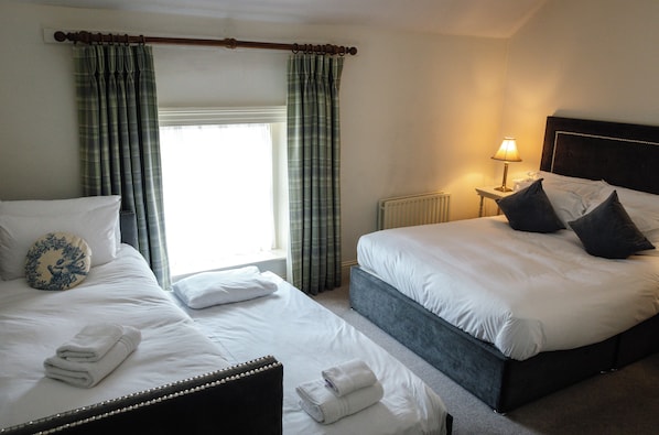 A hotel room with two beds, a window with curtains, a bedside table with a lamp, and a radiator.