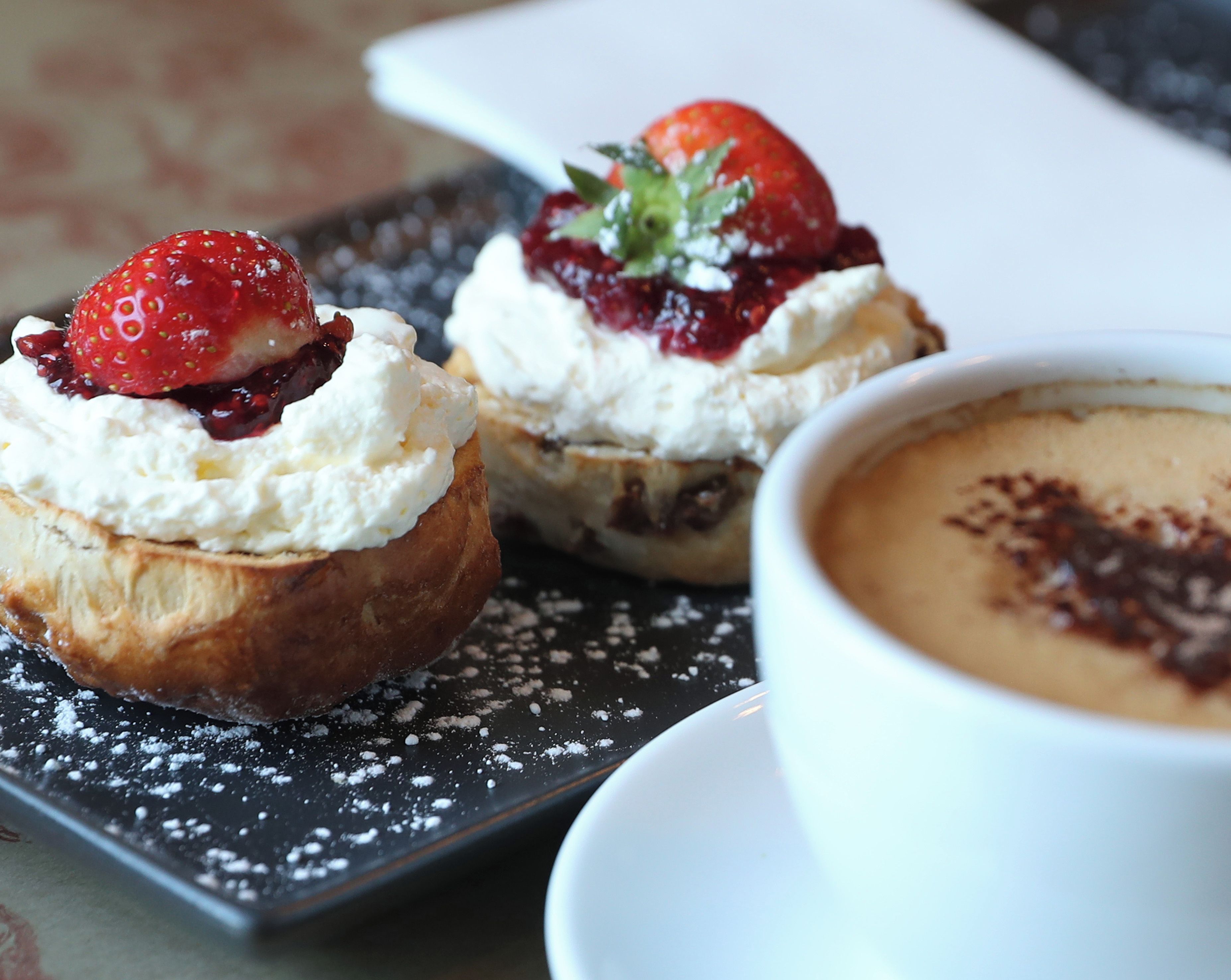 Ein Dessertteller mit zwei Gebäckstücken, bedeckt mit Schlagsahne und Erdbeeren, sowie eine Tasse Kaffee mit Schokoladendrizzle.