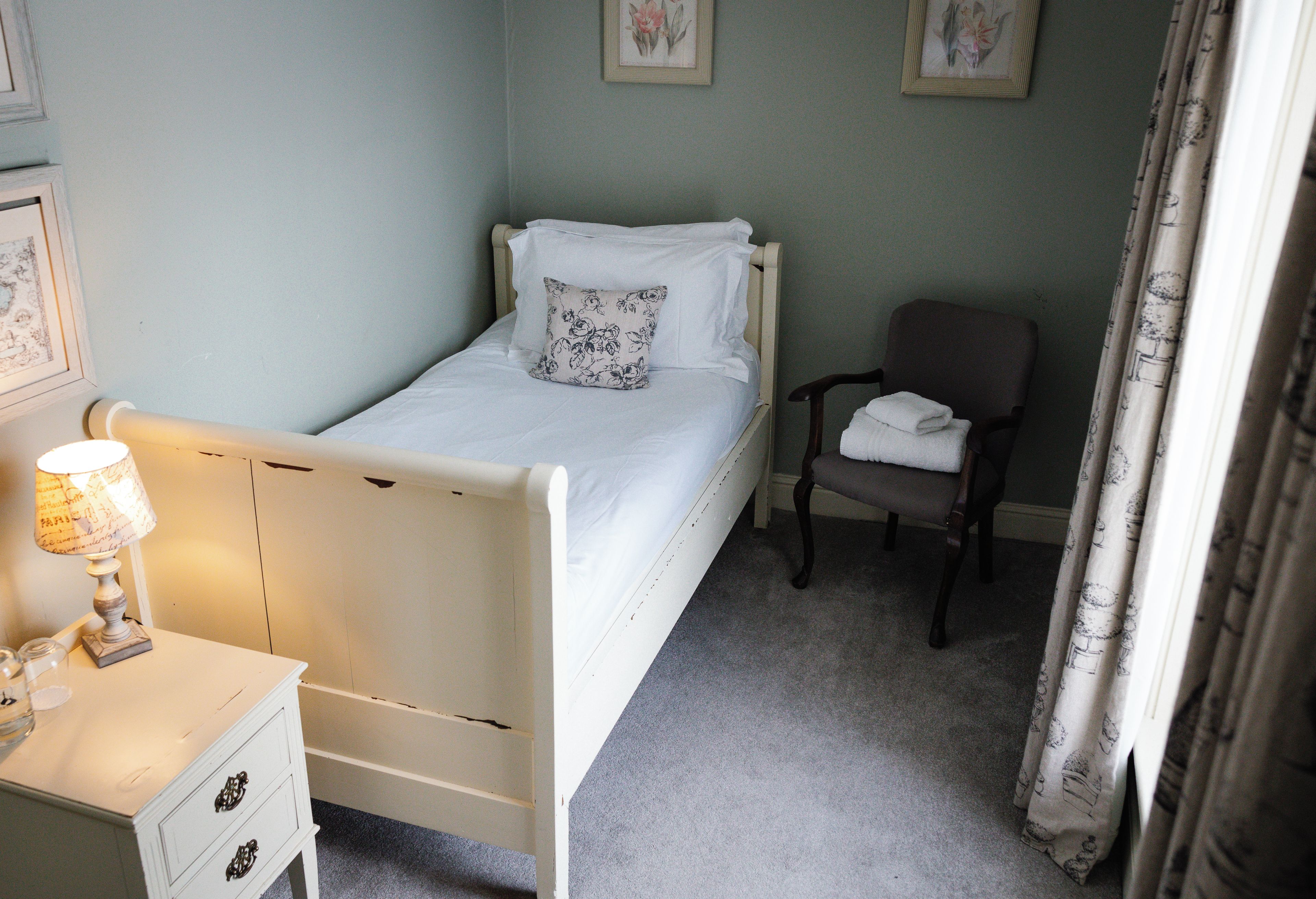 Quarto com uma cama de solteiro, uma cadeira, mesa de cabeceira com abajur e cômoda pequena.