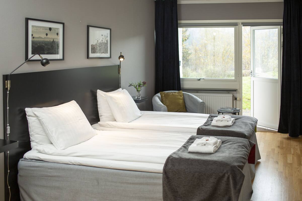 A hotel room with two beds, a chair, a desk, a lamp, and a window with curtains.