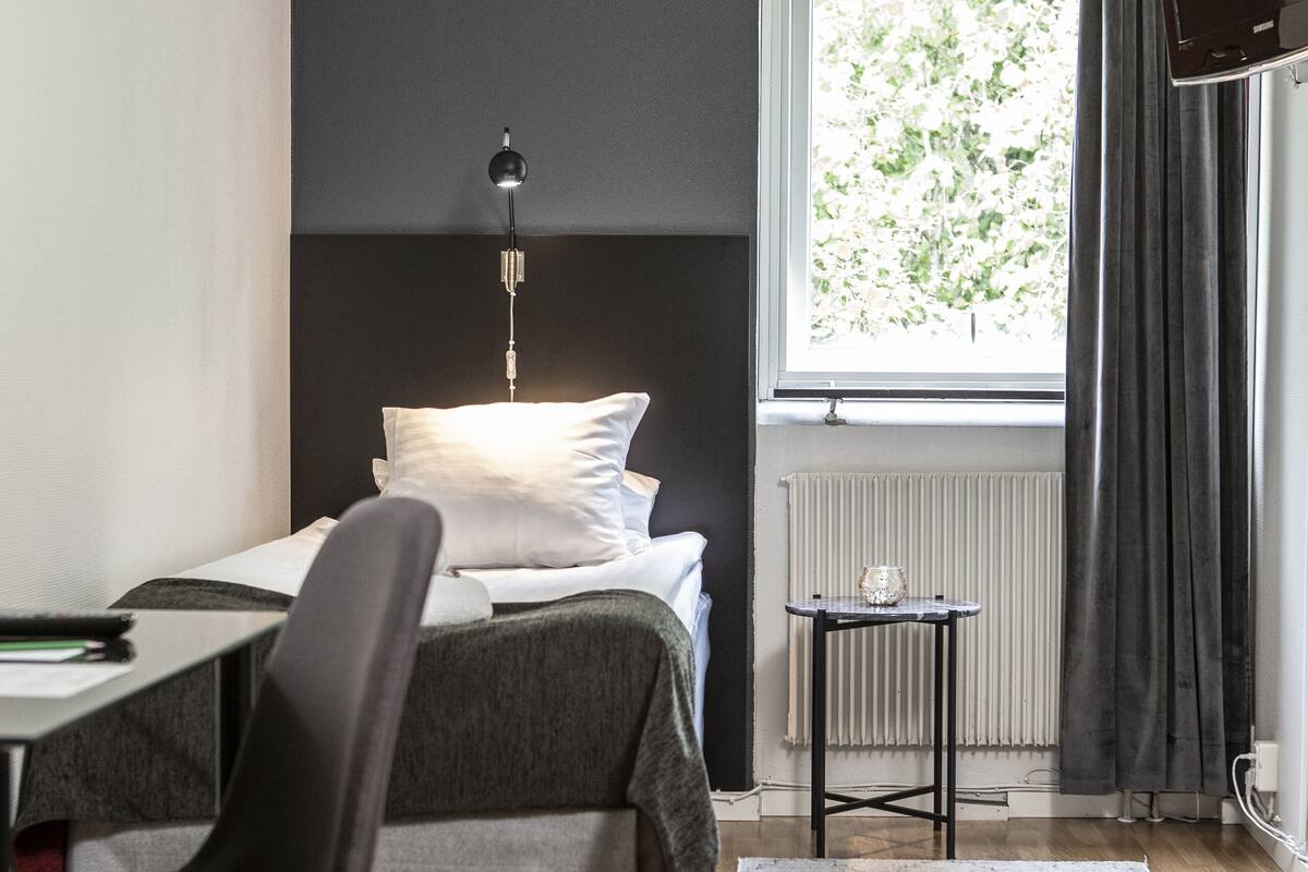 A modern hotel room with a bed, a desk, a chair, a small table, a TV, and a window with curtains.