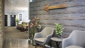 A lobby area with two grey armchairs, a small table, and potted plants. A wooden arrow sign points to the left.