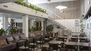A restaurant with a modern interior, featuring a mix of seating options including tables and a sofa area. There is a staircase leading to an upper level, and the space is decorated with plants and framed pictures.