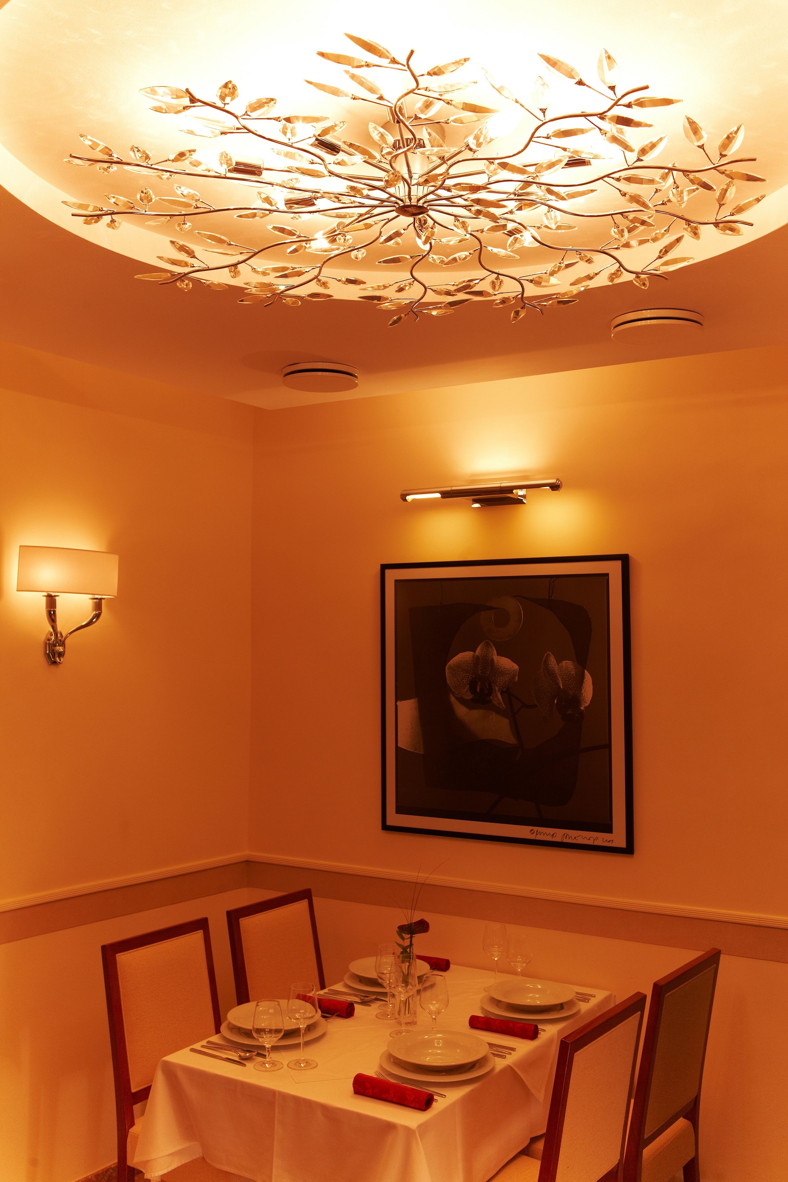 A dining area with a table set for four, a chandelier, wall-mounted lighting, and a framed picture on the wall.