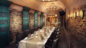 Una larga mesa de comedor cubierta con manteles blancos, velas y cubertería, rodeada de paredes de piedra y sillas de cuero en una habitación tenuemente iluminada.