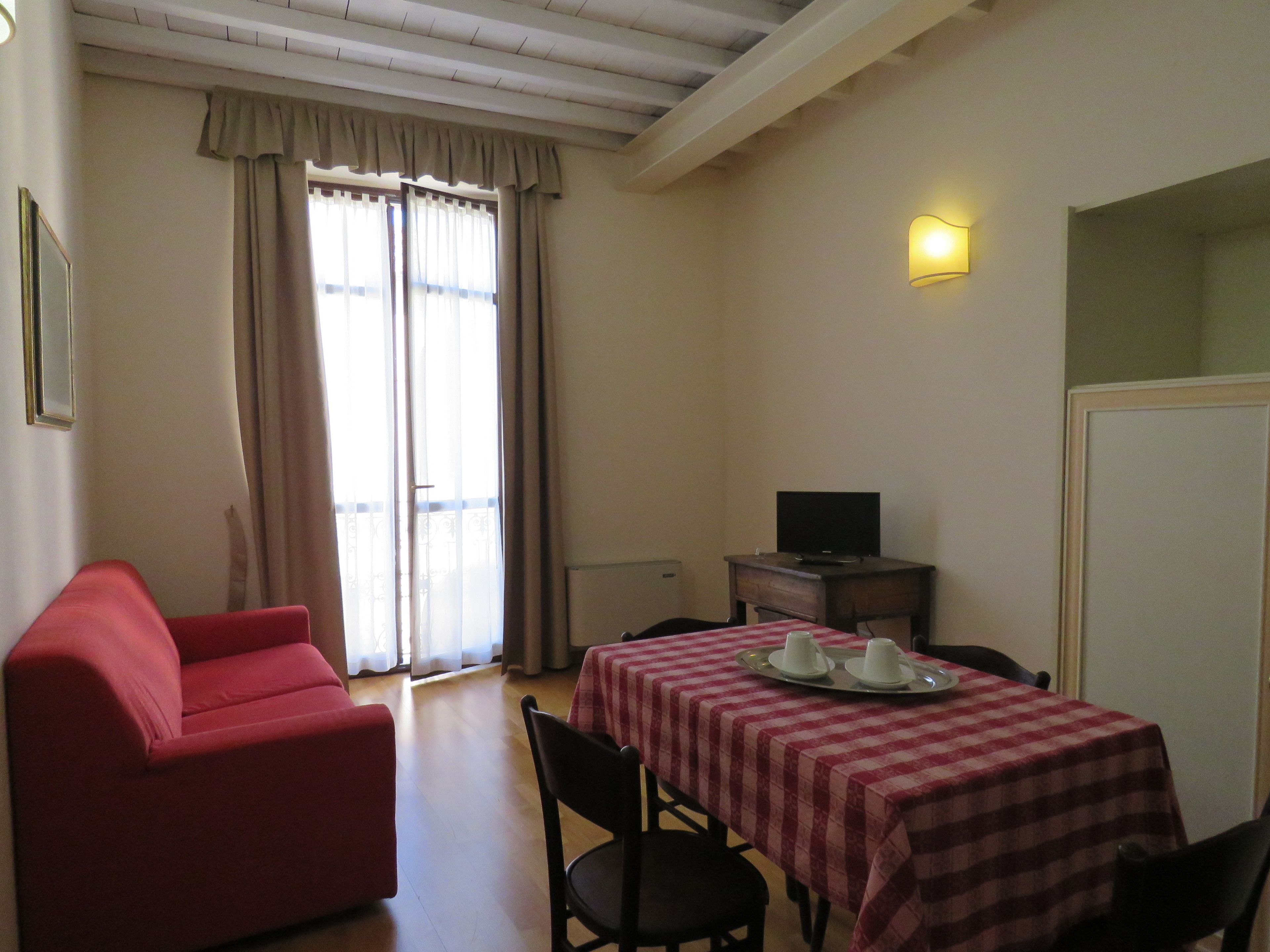 Una habitación con un sillón rojo, una mesa con mantel a cuadros, un televisor sobre una mesita y una ventana con cortinas.