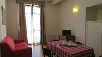 Una habitación con un sillón rojo, una mesa con mantel a cuadros, un televisor sobre una mesita y una ventana con cortinas.