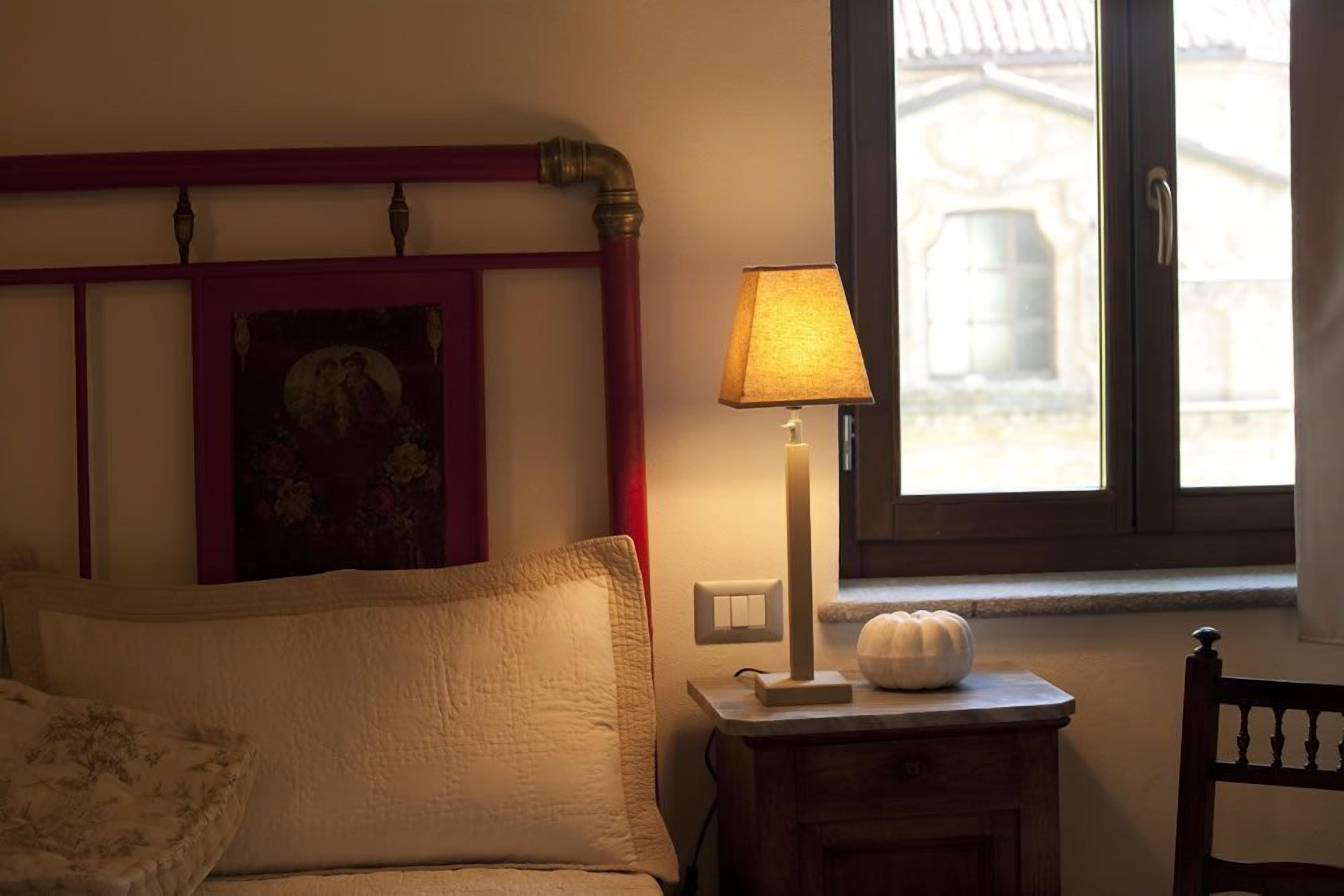 Un dormitorio con cama, una mesita de noche con lámpara, una ventana con vistas a un edificio y un cuadro colgado en la pared.