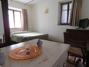 Ein Zimmer mit einem Bett, einem Schreibtisch mit einer Vase, einem Sessel, einem Fernseher und einem Fenster mit Vorhängen.