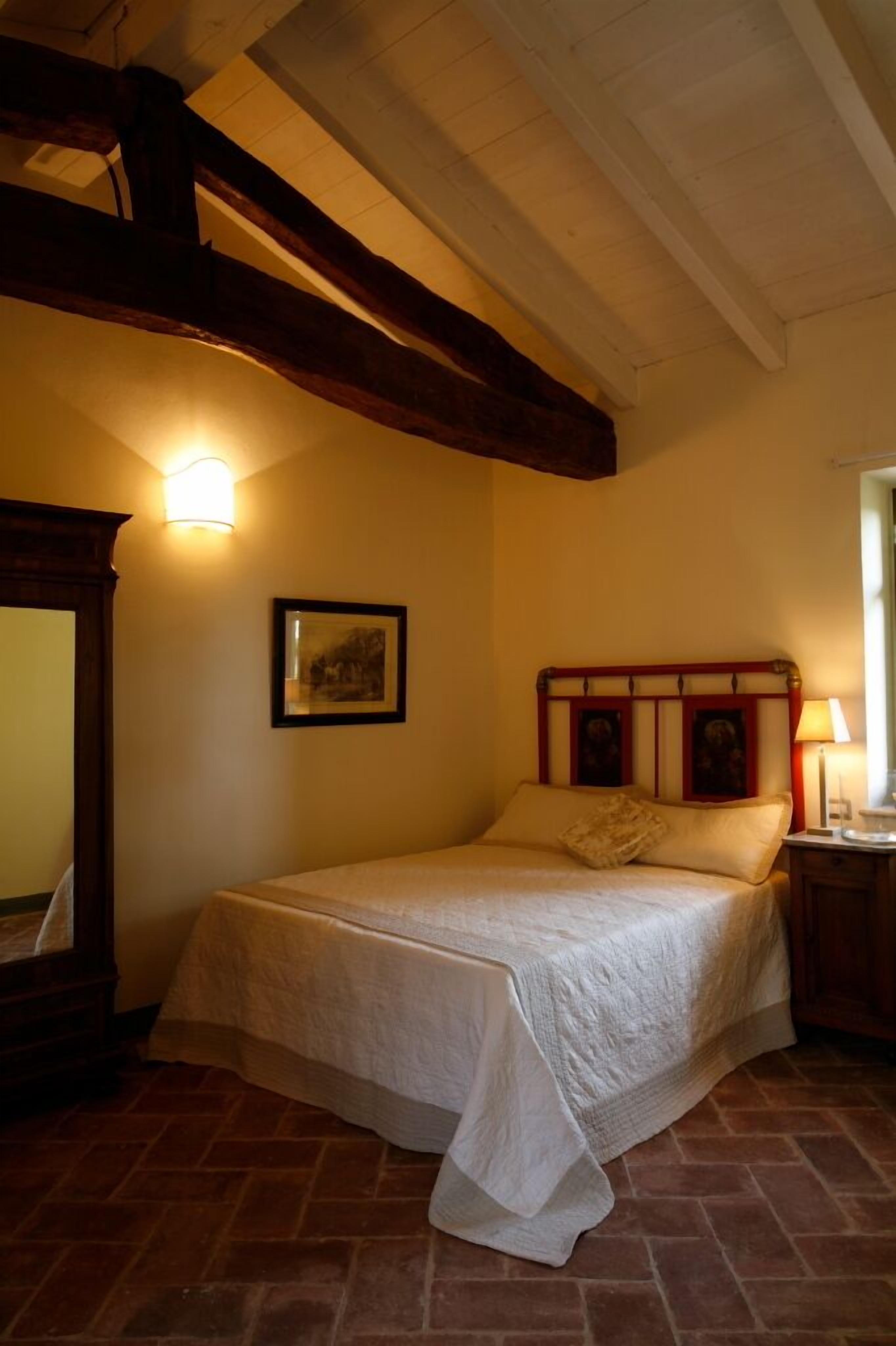 A bedroom with a bed, a nightstand, a framed picture on the wall, and a wooden ceiling beam.