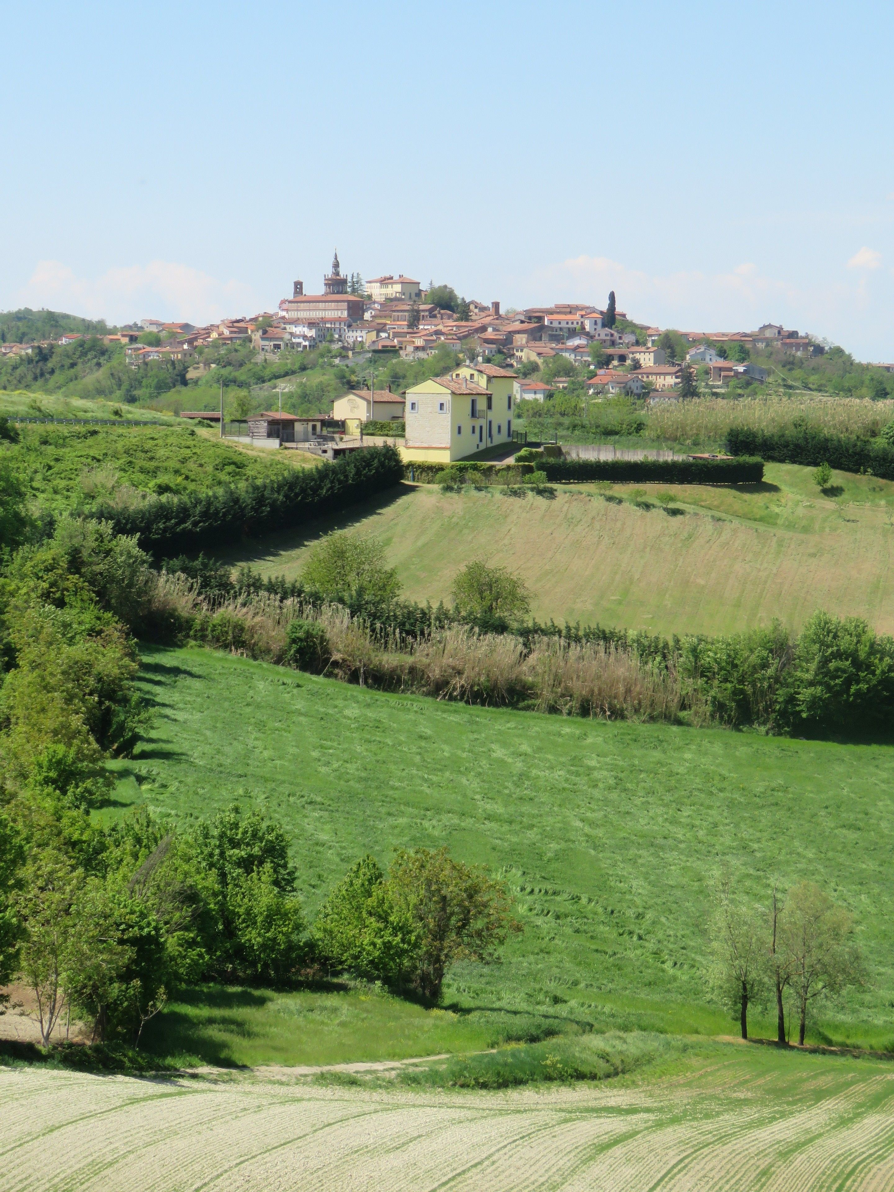 Paesaggio rurale con dolci colline, una piccola casa gialla e un paese in lontananza con edifici dai tetti rossi.