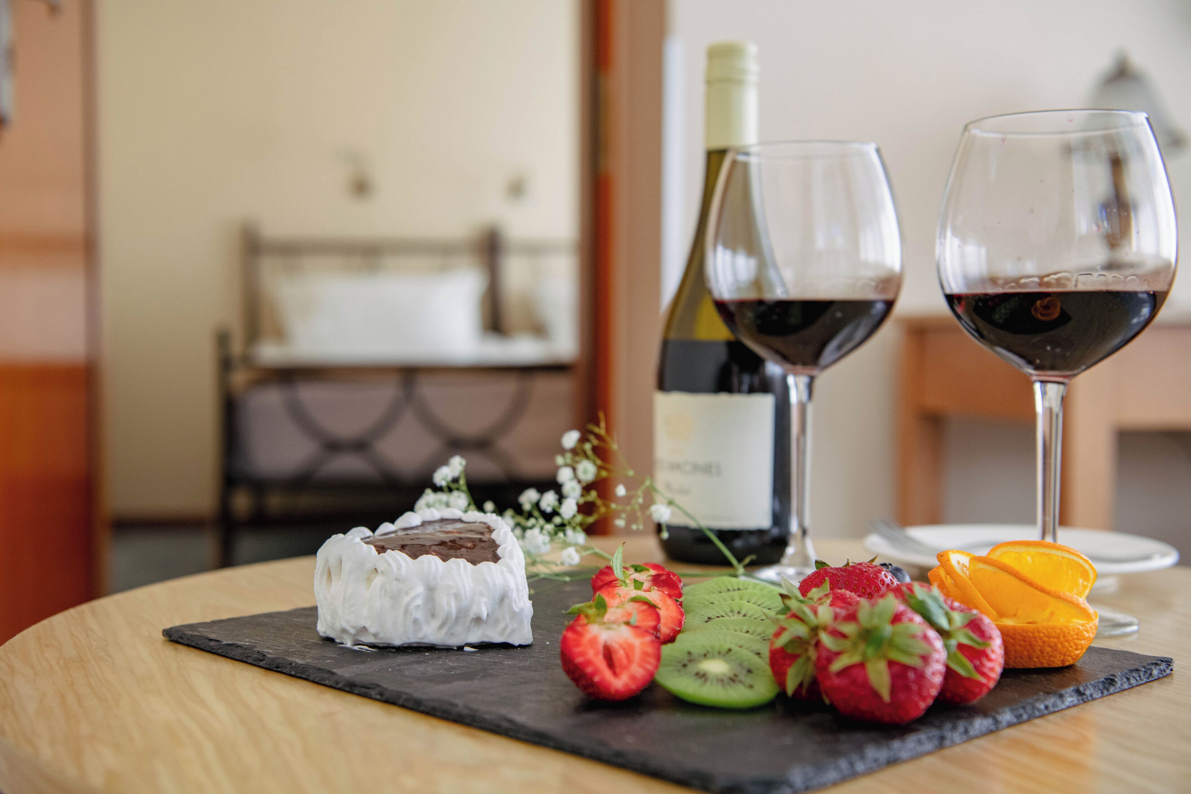 A dessert plate with a chocolate cake, strawberries, kiwi, and orange slices on a slate board, accompanied by two wine glasses and a bottle of wine on a wooden table.