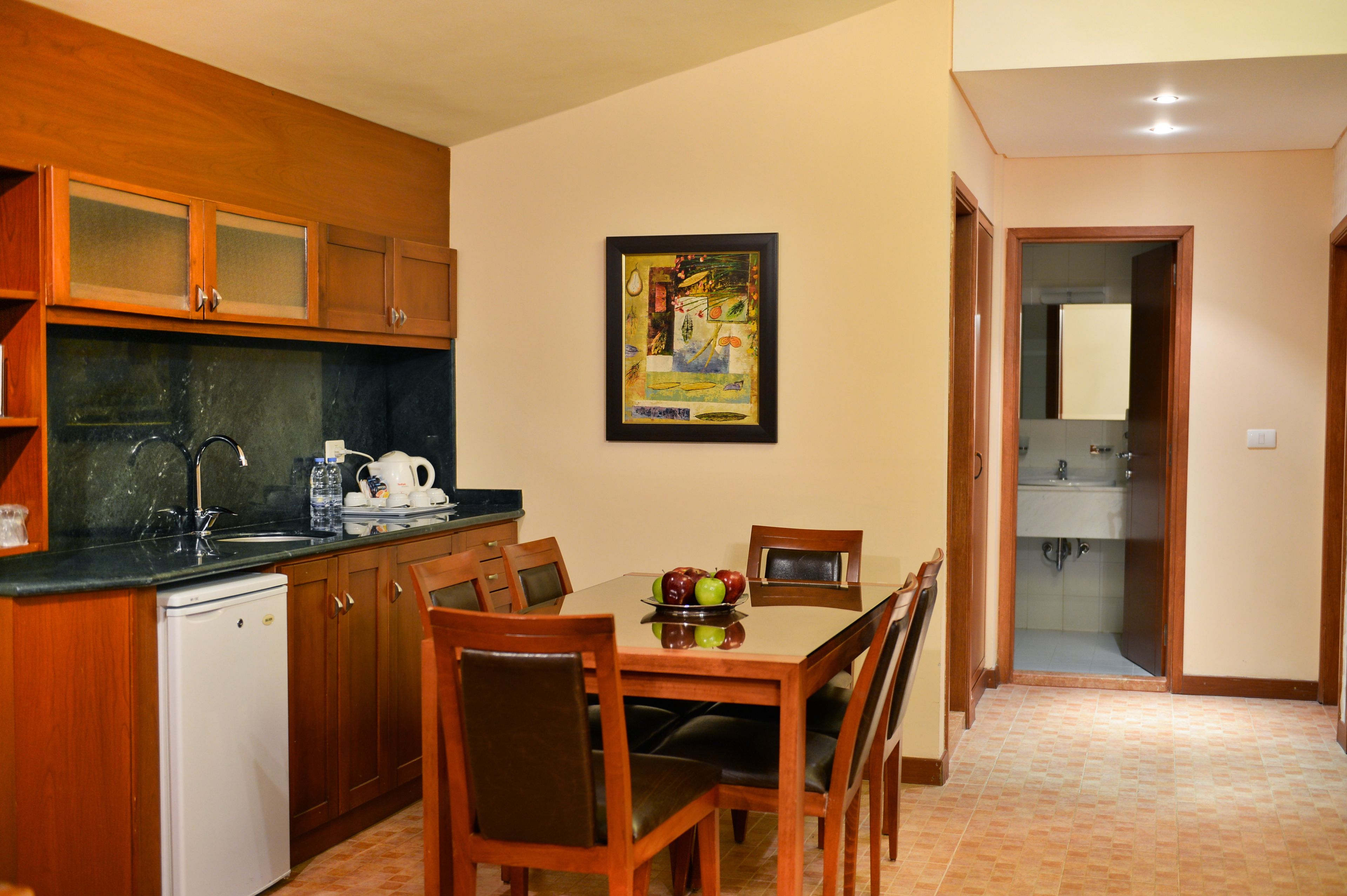 Una cocina compacta con armarios de madera, un refrigerador y una mesa de comedor con sillas. Un cuadro enmarcado adorna la pared, y se ve un lavamanos en el baño contiguo.