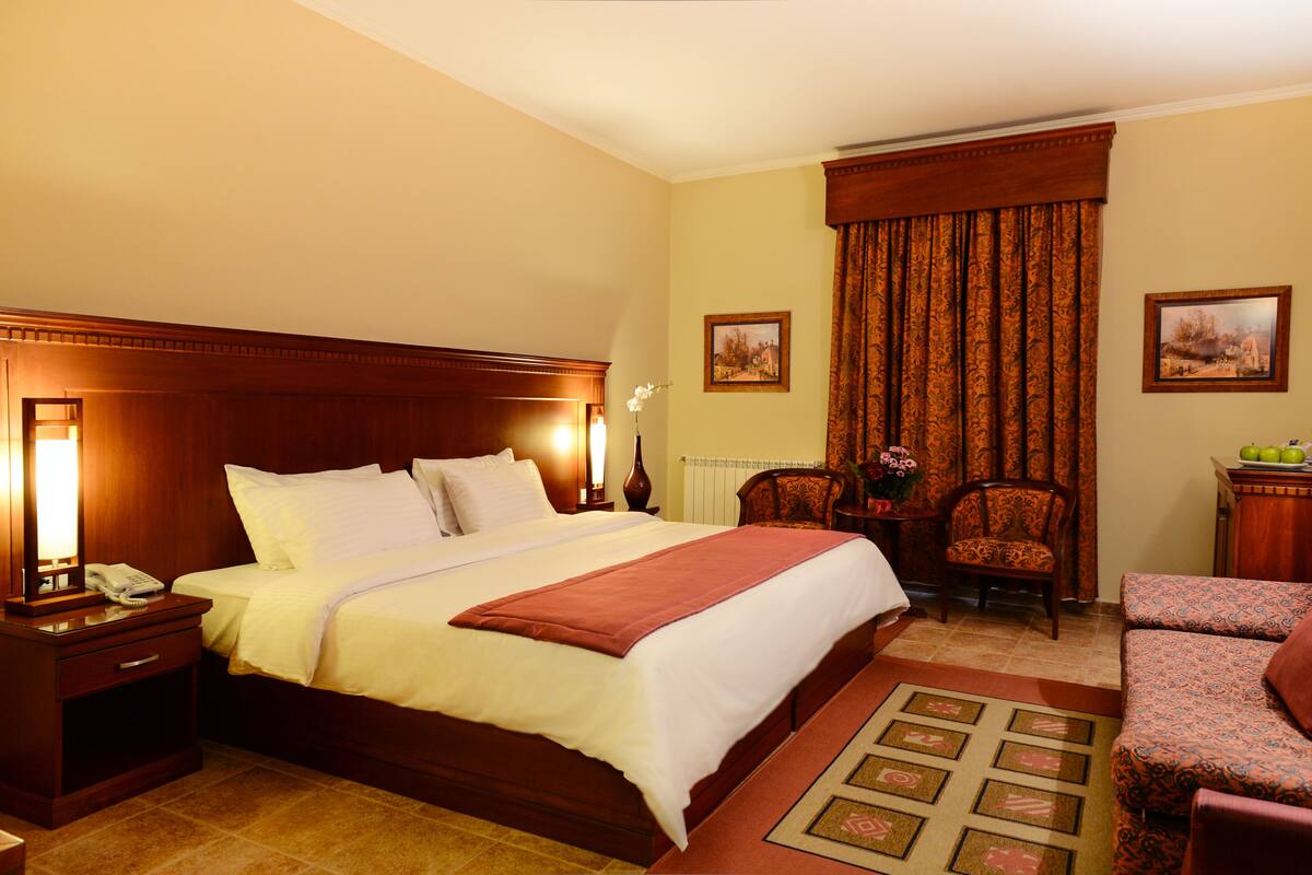 A hotel room with a large bed, a sitting area with a chair, a small table with fruit, and two framed pictures on the wall.